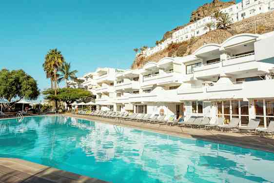 En pool framför ett vitt hotellkomplex med flera balkonger. I bakgrunden syns palmer och en sluttning. (AI-genererad text)