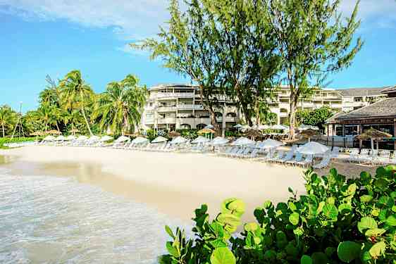 En strand med solstolar och parasoll framför en resort med palmer och grönska. (AI-genererad text)