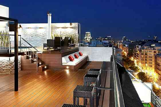 En takterrass med belysning, möbler för utomhusservering och planteringar, med utsikt över en stad på kvällen. (AI-genererad text)