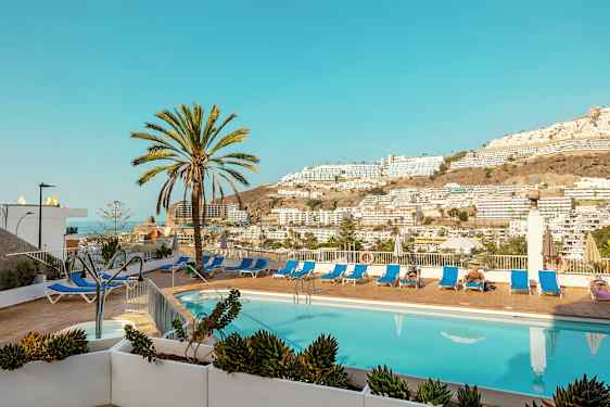 Utomhuspool med solstolar och palmer samt utsikt över berg och byggnader. (AI-genererad text)