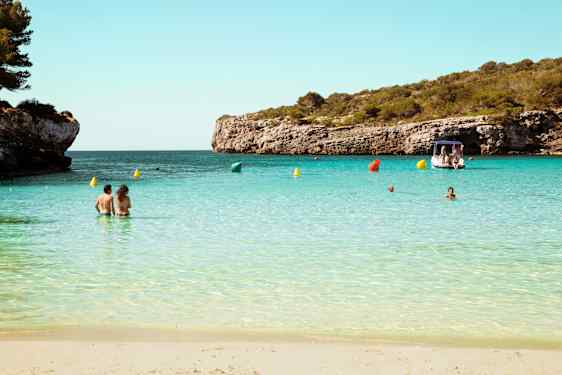 En sandstrand med människor i det lugna, turkosa vattnet. I bakgrunden syns en båt och klippor täckta av grönska. (AI-genererad text)