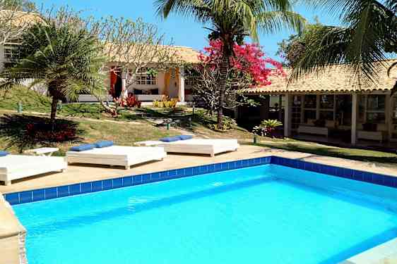 En pool omgiven av solstolar och tropiska växter vid en byggnad med terrakottatak. (AI-genererad text)