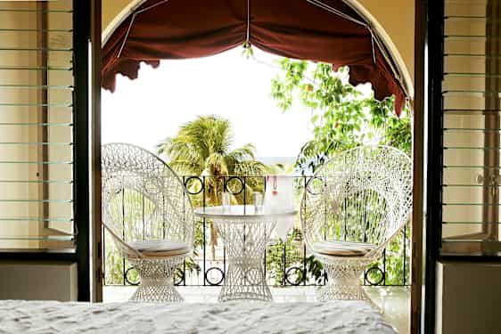 Utsikt från ett rum mot en terrass med två vita stolar och ett bord med glas, omgivna av grönska och palmer. (AI-genererad text)