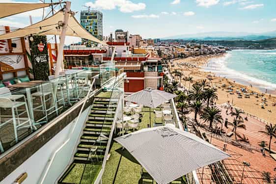 En takterrass med bord och parasoller, omgiven av glasräcken, med utsikt över en strand och staden vid havet. (AI-genererad text)