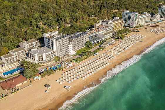 Vy över hotell vid kusten, med strand och parasoller samt grönska i bakgrunden. (AI-genererad text)