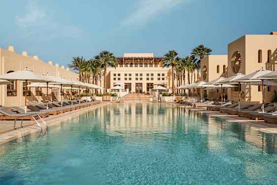 En stor utomhuspool omgiven av solstolar med vita parasoller samt en byggnad i bakgrunden. (AI-genererad text)