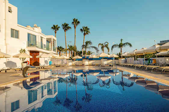 Bild av en utomhuspool omgiven av solstolar och parasoller, med palmer och en vit byggnad i bakgrunden. (AI-genererad text)
