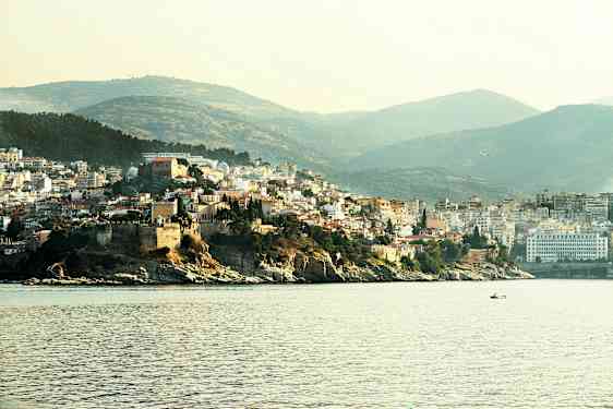 Bild som visar en kuststad med bebyggelse på en höjd nära havet, med båtar på vattnet och gröna kullar i bakgrunden. (AI-genererad text)