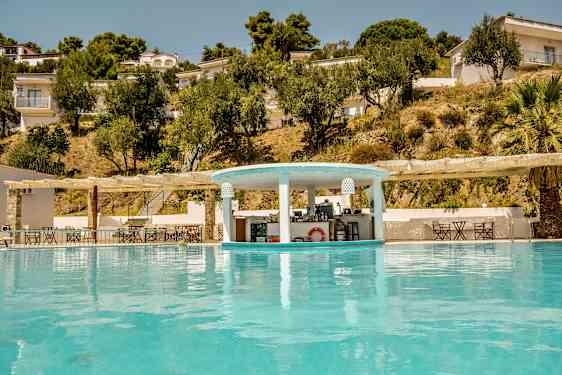 En pool med en poolbar och omgivande byggnader och träd på sluttningen. (AI-genererad text)