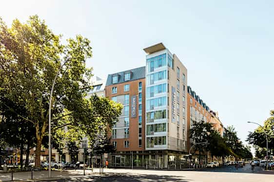 Ett modernt hotell med glasfasad omgiven av träd och stadsmiljö. (AI-genererad text)