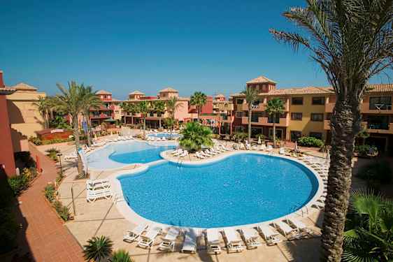 Hotellområde med pool och solstolar, omgiven av palmer och byggnader i varma färger. (AI-genererad text)