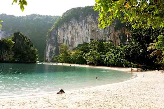Sandstrand med klart vatten, omgiven av gröna träd och höga kalkstensklippor. (AI-genererad text)