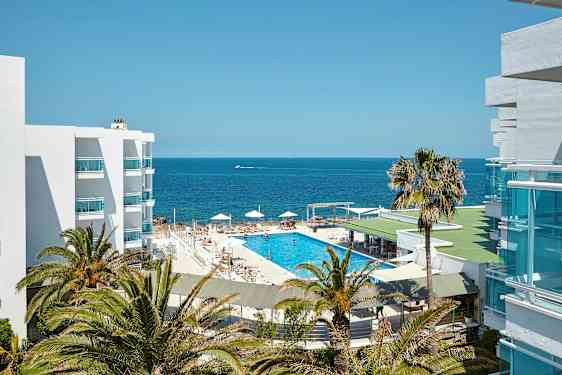 En bild av ett hotellområde med utsikt över havet. Det finns en swimmingpool omringad av solstolar och parasoller, omgiven av palmer och vita byggnader. (AI-genererad text)