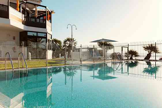 En pool omgiven av ett glasstaket med utsikt mot en gräsmatta och solstolar under parasoll. (AI-genererad text)