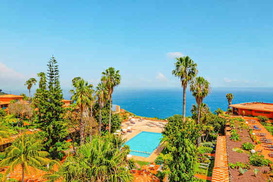 Utsikt över ett poolområde omgivet av grönska och palmer, med havet i bakgrunden. (AI-genererad text)