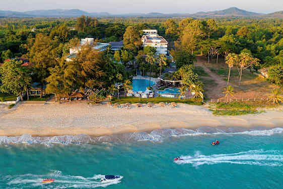 En strand med parasoller och stolar samt ett hotellkomplex och pooler i bakgrunden, omgiven av växtlighet. (AI-genererad text)