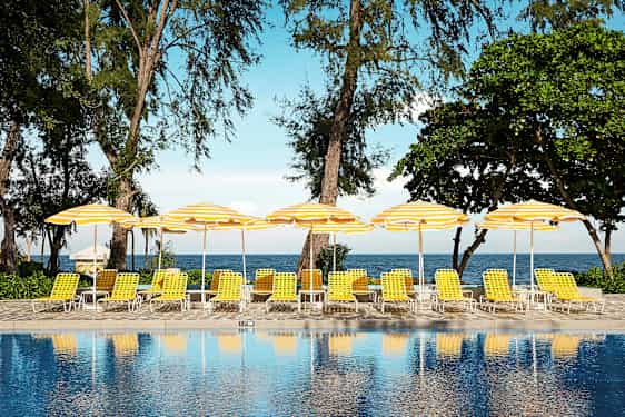 En pool framför solstolar med gula och vita parasoller nära stranden. (AI-genererad text)