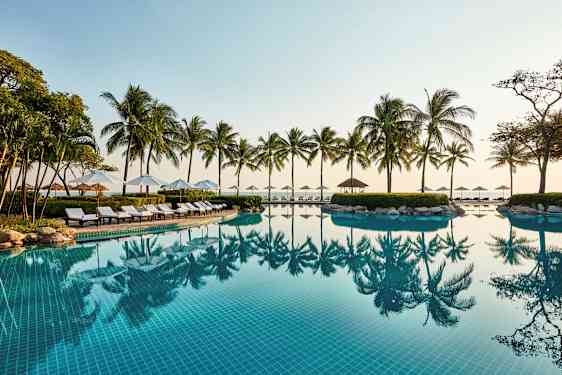 En utomhuspool med klarblått vatten, omgiven av solstolar, parasoller och palmer vid havet. (AI-genererad text)