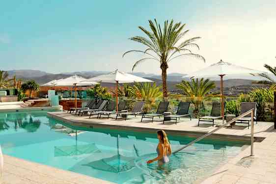 Poolområde med solstolar och parasoller, omgiven av palmer och med utsikt över landskap. (AI-genererad text)