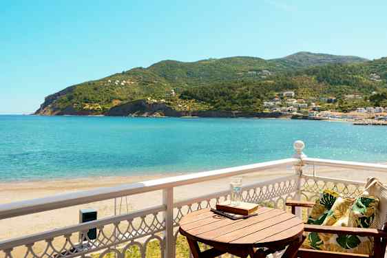 Utsikt från en balkong mot havet och grönskande kullar, med en trästol och ett bord. (AI-genererad text)