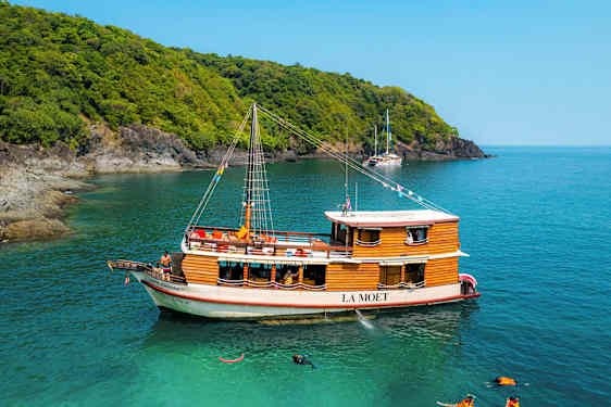 En träbåt med namnet La Moet är förtöjd vid turkost vatten nära en skogsklädd kust. (AI-genererad text)