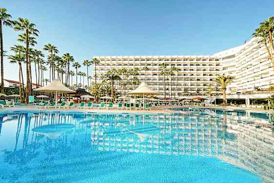 Hotellbyggnad med stor pool och solstolar i förgrunden, omgiven av palmer. (AI-genererad text)