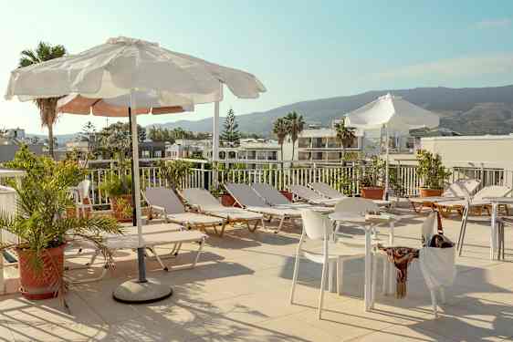 En solterrass med solstolar, parasoller och bord samt stolar omgivna av grönska och med utsikt över byggnader och berg. (AI-genererad text)