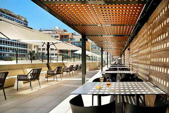 En utomhusterrass med bord och stolar under ett träpergola och stora parasoller. (AI-genererad text)