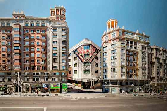 Tre byggnader ligger längs en gata, där mittbyggnaden har färgglada konstverk på fasaden. (AI-genererad text)