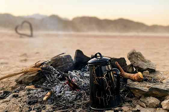 En kaffekanna står vid en slocknad lägereld i en ökenlandskap. (AI-genererad text)