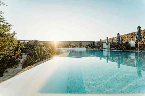 En swimmingpool med parasoller och solstolar, omgiven av växter och stenmur, med solen i bakgrunden. (AI-genererad text)