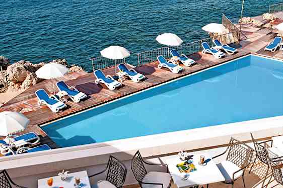 En pool med solstolar och parasoller vid en träterrass intill havet. Förgrunden visar ett bord med stolar och drycker. (AI-genererad text)