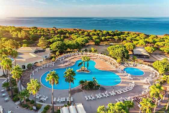Flygfoto av ett hotellområde med stora pooler, solstolar och omgivande grönska nära stranden. (AI-genererad text)