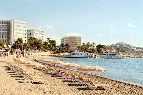 Strand med solstolar och parasoller, hotellbyggnader i bakgrunden och båtar på vattnet. (AI-genererad text)
