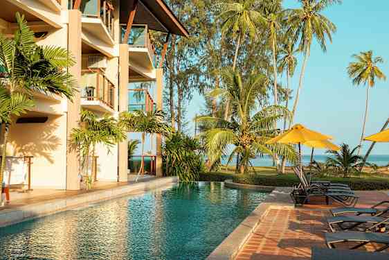 En utomhuspool bredvid ett hotell med balkonger, omgiven av tropiska palmer och gräsyta vid havet. (AI-genererad text)