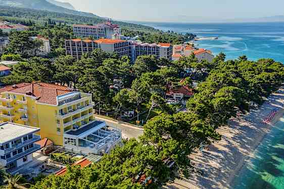 Luftvy över en strand med solstolar och parasoller bredvid hotellbyggnader och grönska. (AI-genererad text)