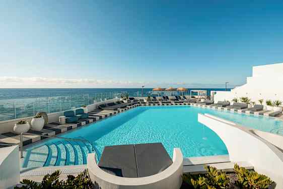 En pool med turkost vatten omgivet av solstolar och utsikt över havet. I bakgrunden syns en klar blå himmel. (AI-genererad text)