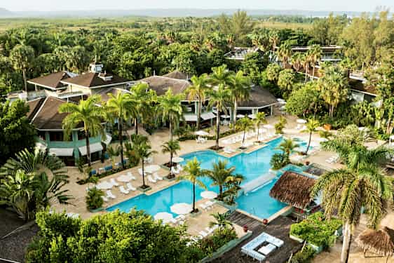 Flygfoto över ett hotellområde med en stor pool omgiven av solstolar och parasoller, samt grönskande träd och vegetation. (AI-genererad text)