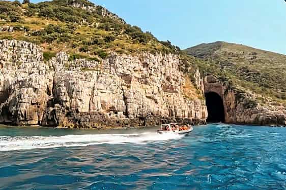 En motorbåt med människor färdas på blått vatten nära en klippa med en stor grottöppning. (AI-genererad text)