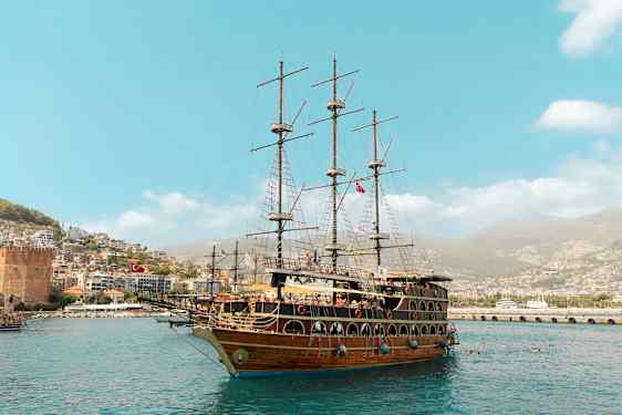 Ett stort segelfartyg ligger i hamninloppet, med en stad och gröna kullar i bakgrunden. (AI-genererad text)