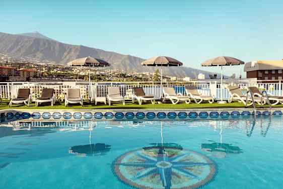 Poolområde med solstolar och parasoller med utsikt över berg och byggnader. (AI-genererad text)