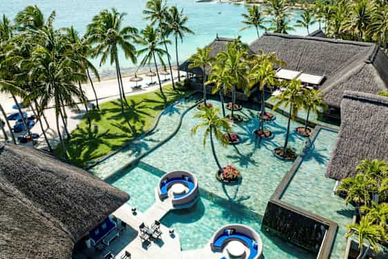 Flera hus med tak av halm vid en pool med palmer och sandstrand i bakgrunden. (AI-genererad text)