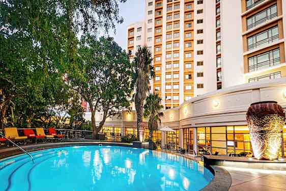 En upplyst utomhuspool omgiven av gröna växter och en byggnad i bakgrunden. (AI-genererad text)