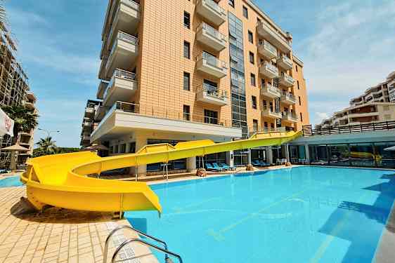 En byggnad med balkonger bredvid en pool med en gul vattenrutschkana. (AI-genererad text)
