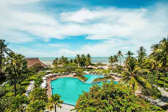 En utomhuspool omgiven av gröna träd och parasoller med utsikt mot havet under en klar blå himmel. (AI-genererad text)