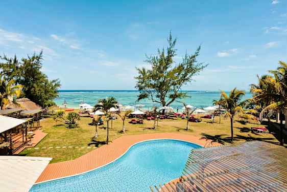 En pool med turkost vatten framför en gräsmatta med träd, solsängar och parasoller längs en strand. (AI-genererad text)