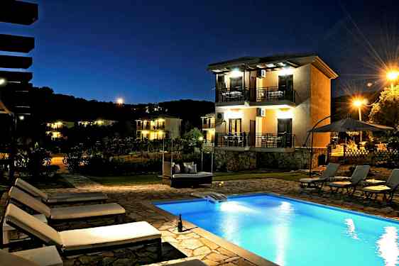 En kvällsbild av en utomhuspool och flera solstolar. Bakgrunden visar ett hus med balkonger och omgivande belysning. (AI-genererad text)