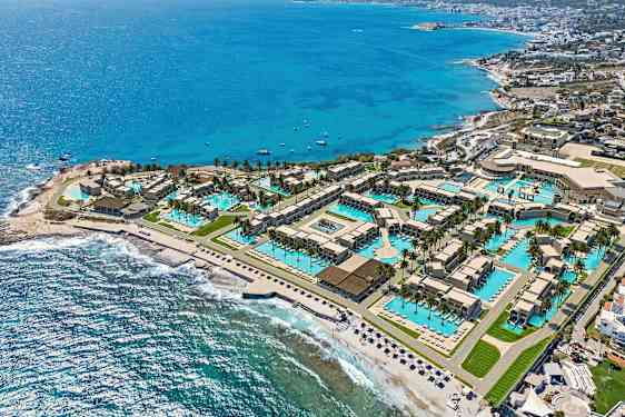 Flygbild över en semesterort vid kusten med pooler, byggnader och stranden intill havet. (AI-genererad text)