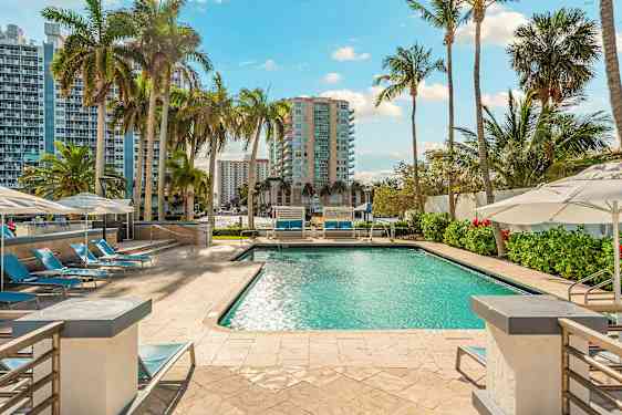 En swimmingpool omgiven av solstolar och parasoller, med höga byggnader och palmträd i bakgrunden. (AI-genererad text)