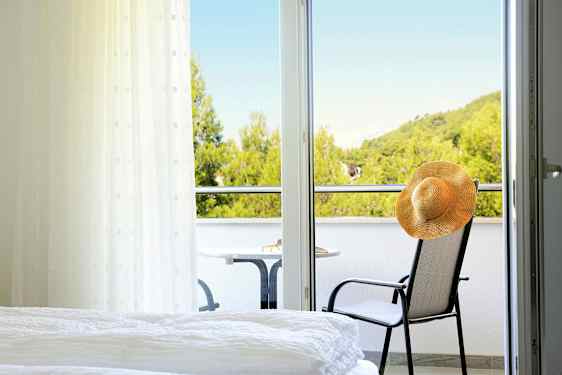 Ett hotellrum med utsikt över en balkong där en stol med en hatt och ett bord står, samt grönskande träd i bakgrunden. (AI-genererad text)
